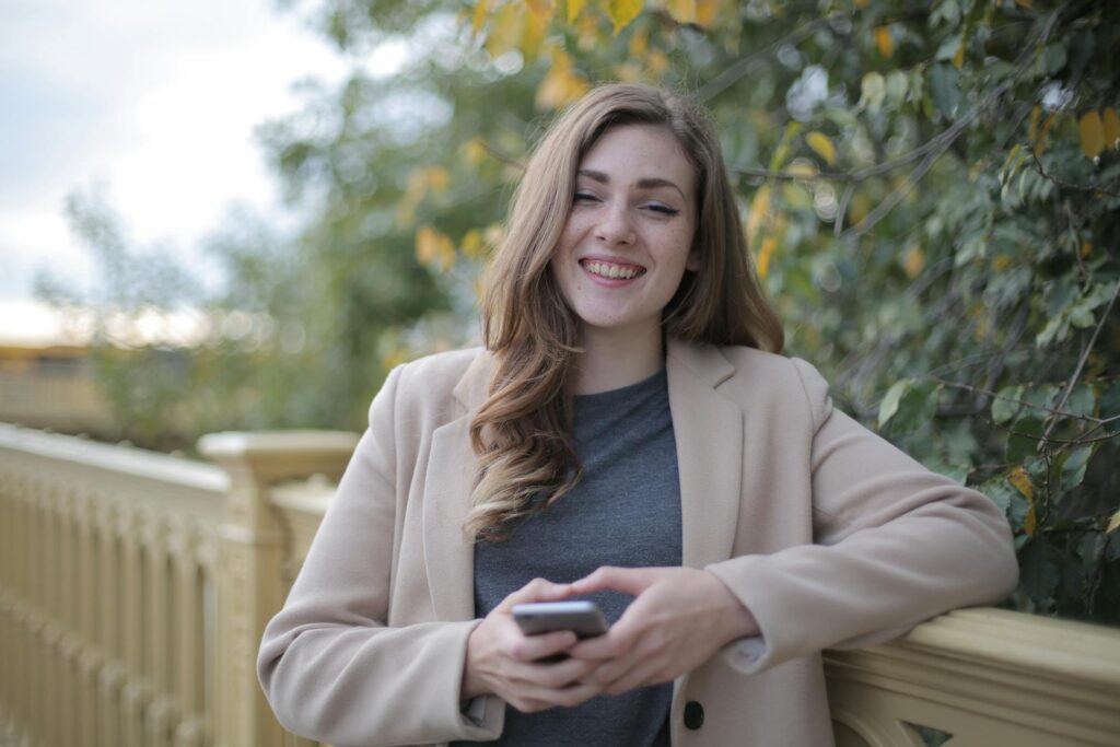 A happy woman holding a smartphone leading against a railing.